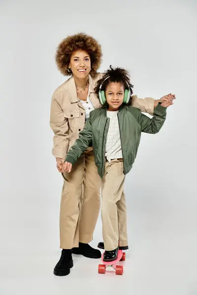Stylish curly African American mother and daughter posing gracefully on a skateboard against a grey background. — Stock Photo