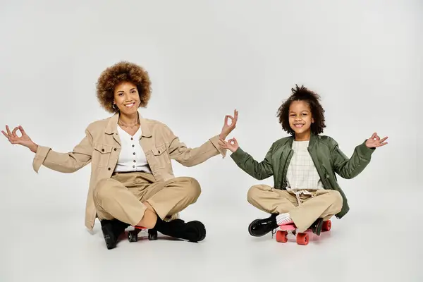 Curly African American mother and daughter in stylish outfits, sitting on skateboards, practicing yoga together on a grey background. — Stock Photo