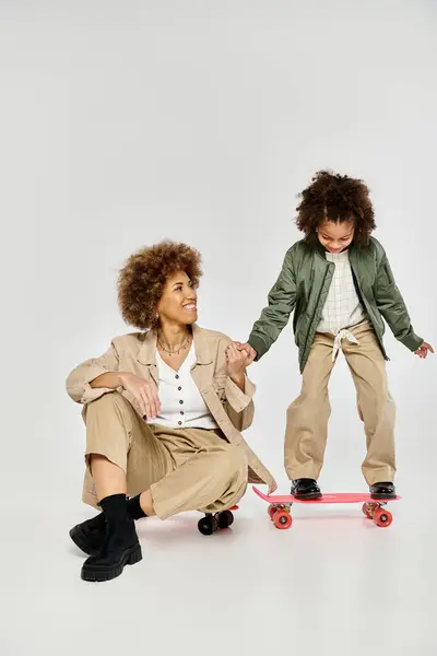 A curly African American mother and daughter in stylish clothes enjoy skateboarding together on a grey background. — Stock Photo