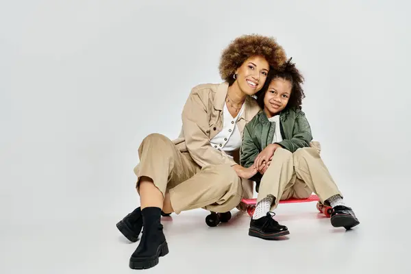 Curly African American mother and daughter posing stylishly on a skateboard against a grey background. — Stock Photo