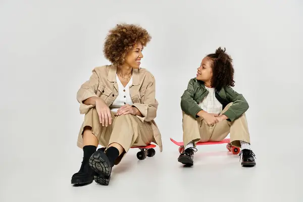 Curly mãe e filha afro-americana sentar elegantemente em um skate contra um pano de fundo cinza. — Fotografia de Stock