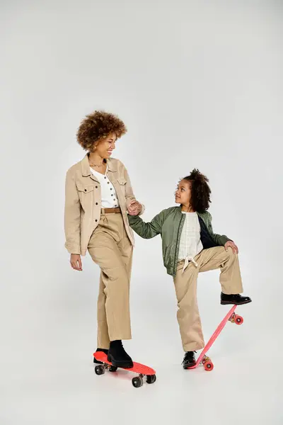 A curly African American mother and daughter, both in stylish clothes, standing confidently on a skateboard against a grey background. — Stock Photo