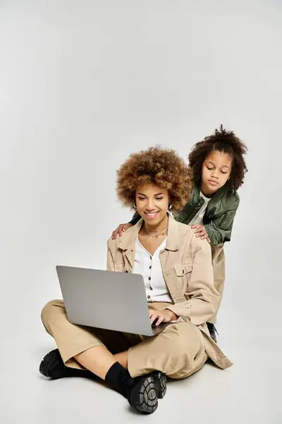 Curly mère afro-américaine et sa fille assis sur le sol, en utilisant un ordinateur portable ensemble dans des vêtements élégants. — Photo de stock
