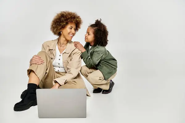 Curly African American mother and daughter in stylish clothes, sitting on the floor, focused on a laptop screen. — Stock Photo