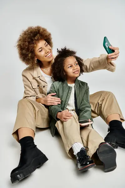 Curly African American mother and daughter in stylish clothes taking a selfie with a cell phone on a grey background. — Stock Photo