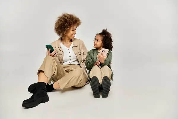 Ricci afroamericani madre e figlia in abiti eleganti seduti sul pavimento, assorti in un telefono cellulare. — Foto stock