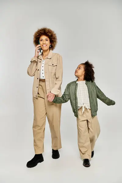 La madre y la hija afroamericanas rizadas con ropa elegante entablan una conversación telefónica sobre un fondo gris. - foto de stock