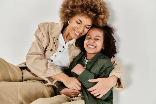 A curly African American mother hugs her daughter affectionately on the floor in stylish clothes against a grey background. — Stock Photo