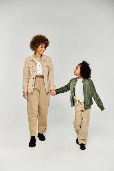 Curly African American mother and daughter holding hands in stylish outfits against a grey studio backdrop. — Stock Photo