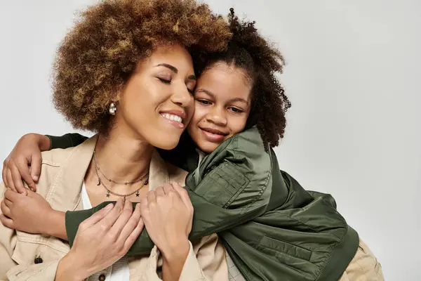 Madre e hija afroamericana rizada con ropa elegante abrazándose ferozmente frente a un fondo blanco. - foto de stock