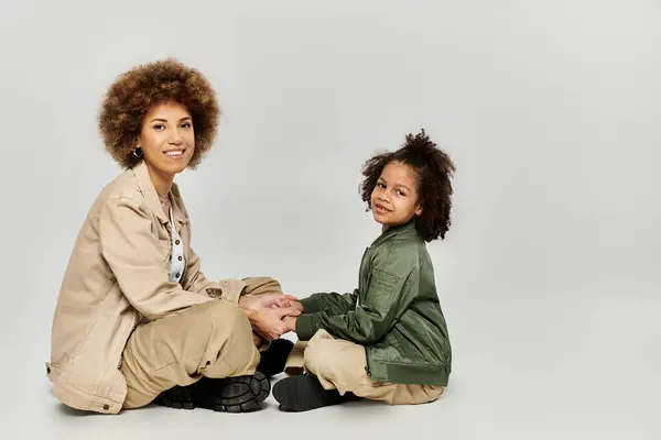 Ricci afroamericani madre e figlia seduti insieme sul pavimento grigio in abiti eleganti, condividendo un momento tenero. — Foto stock