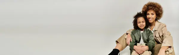 A curly African American mother and daughter in stylish clothes sit together on a white background, sharing a moment of closeness. — Stock Photo