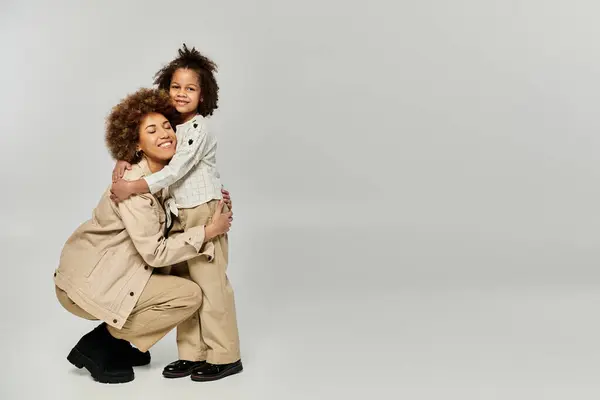 A curly African American mother warmly embraces her daughter, both in stylish attire, against a serene gray background. — Stock Photo
