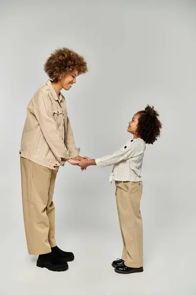 Uma mãe e filha afro-americana encaracolada, vestida com roupas elegantes, ficam ao lado umas das outras em um fundo cinza. — Fotografia de Stock