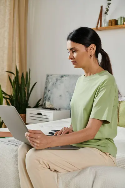 A woman in casual attire sits on a bed, working on a laptop computer. — Stock Photo