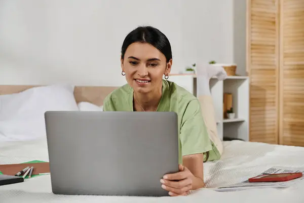 Una mujer con atuendo casual yace en una cama, absorta en su computadora portátil. - foto de stock