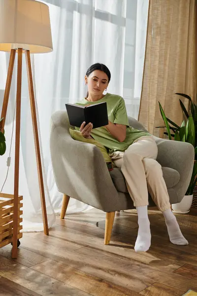 Una mujer en atuendo casual, inmersa en un libro mientras está sentada en una silla en casa. - foto de stock