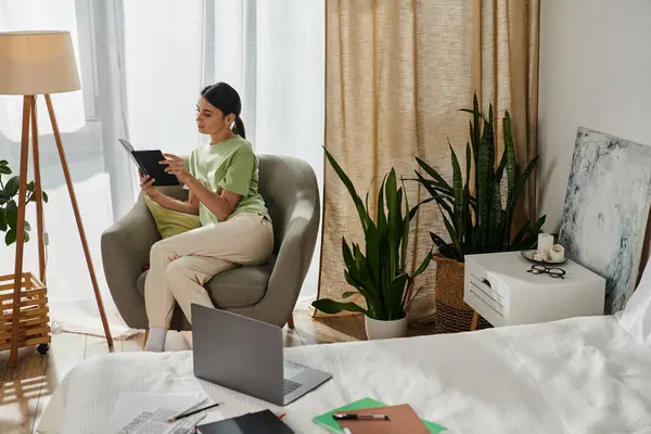 Женщина в повседневной одежде читает книгу рядом с ноутбуком дома. — стоковое фото