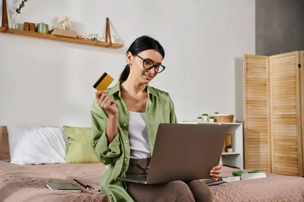 Woman sitting on a bed, holding a credit card and laptop, working remotely. — Stock Photo