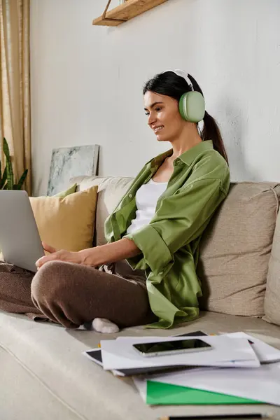 Una mujer con atuendo casual, trabajando desde casa, se sienta en un sofá escribiendo en un portátil. - foto de stock
