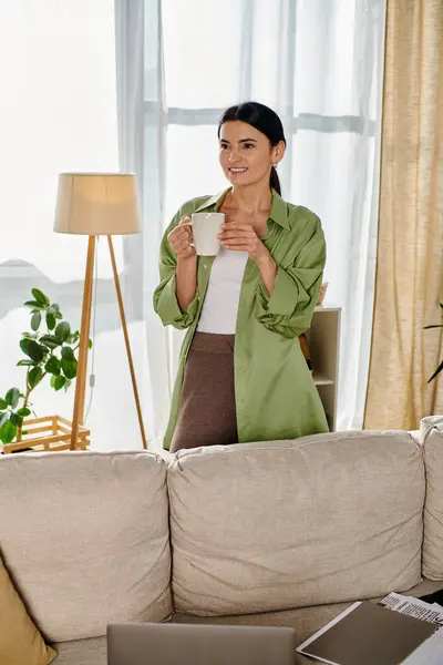 Una donna in abbigliamento casual gode di una tazza di caffè mentre si trova vicino a un divano. — Foto stock