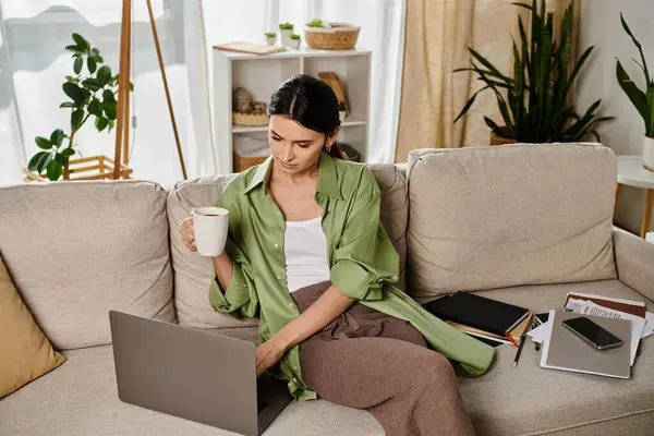 Donna in abbigliamento casual, lavorando su laptop con caffè in mano su un divano accogliente. — Foto stock
