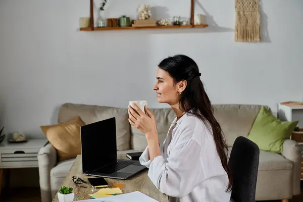 A woman in casual attire sitting at a desk, savoring a cup of coffee. — Stock Photo