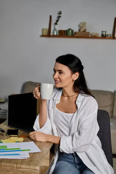 A stylish woman sitting at a table, savoring a cup of coffee while working from home. — Stock Photo