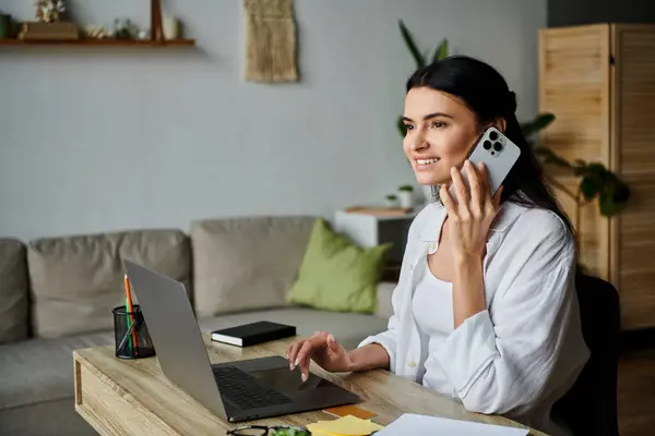 A woman sits at a table, talking on a cell phone while working from home. — Stock Photo
