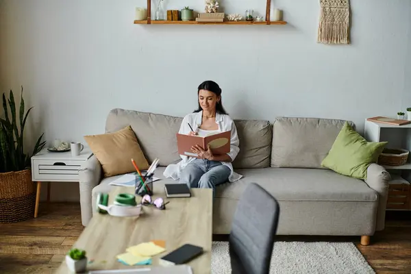 A woman in casual attire sits on a couch, engrossed in a book. — Stock Photo