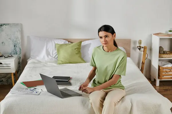 A woman in casual attire sits on a bed, typing on a laptop. — Stock Photo