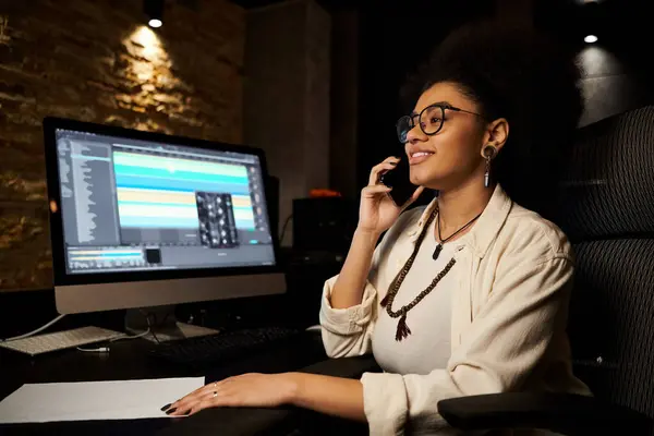 Una mujer multitarea, hablando por teléfono y trabajando en una computadora durante un ensayo de una banda de música en un estudio de grabación. - foto de stock