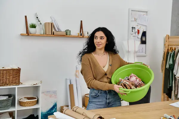 Una joven vestida de manera casual, con la ropa puesta, sosteniendo un lavabo verde lleno de prendas. - foto de stock