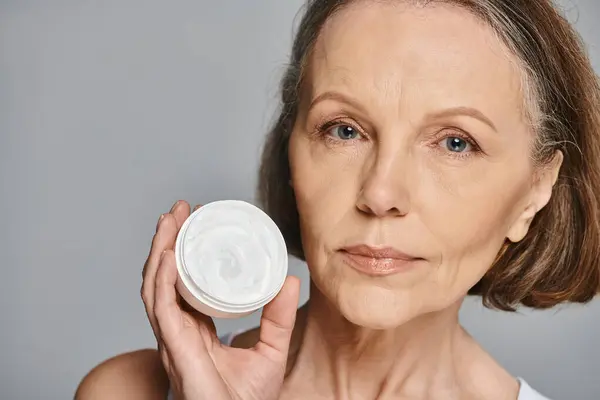 A mature woman in comfortable attire elegantly holding a container of cream. — Stock Photo