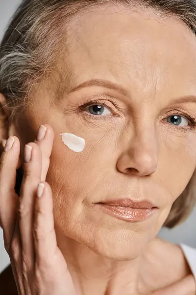 Un'elegante donna anziana che si applica delicatamente la crema sul viso, prendendosi cura della sua pelle. — Foto stock
