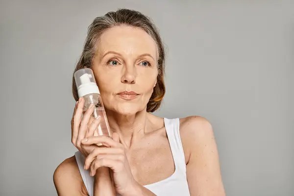 A mature woman in cozy attire holding a facial cleanser bottle. — Stock Photo