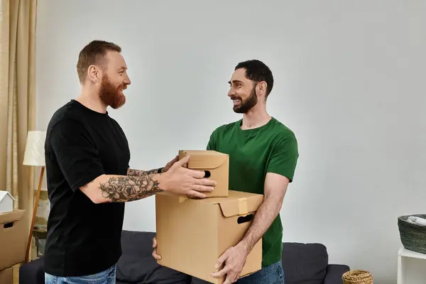 Um casal gay apaixonado está em sua sala de estar, segurando uma caixa de papelão, pronto para começar sua nova vida juntos. — Fotografia de Stock