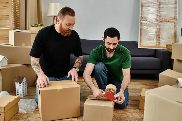 Two men sit on the floor surrounded by moving boxes, embracing the start of a new life in their new home. — Stock Photo
