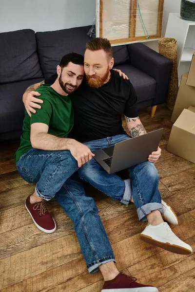 Gay couple in love sits on floor, surrounded by boxes, using laptop in their new home. — Stock Photo