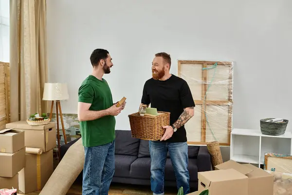 Um casal gay abraça em sua nova sala de estar, segurando uma cesta, cercado por caixas móveis, começando sua nova vida juntos. — Fotografia de Stock
