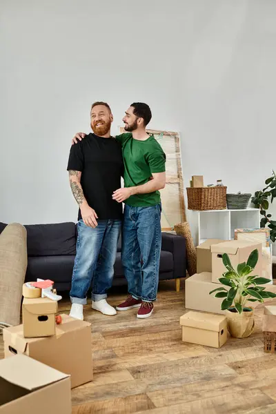 Un couple gay amoureux, entouré de boîtes mobiles, debout ensemble dans leur nouveau salon. — Photo de stock
