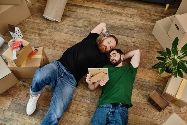 Un couple d'hommes allongés paisiblement sur un plancher de bois dans leur nouvelle maison, un moment de repos au milieu des boîtes mobiles. — Photo de stock
