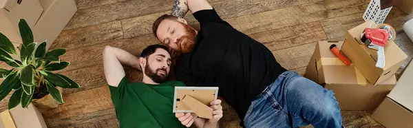 Two men in love lay peacefully atop a wooden floor, beginning their life together in a new home filled with moving boxes. — Stock Photo