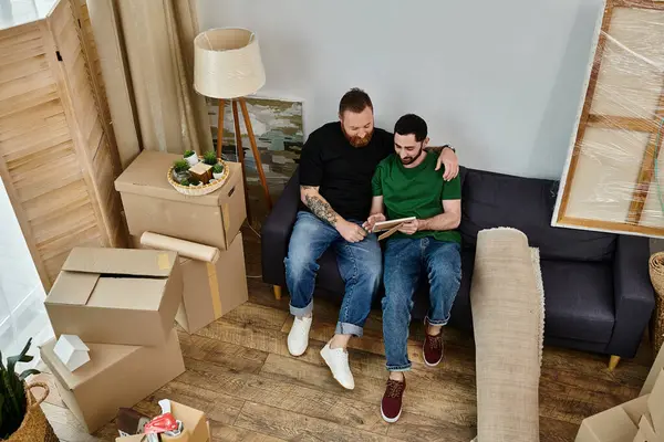 A gay couple sit atop a couch, embracing in their new home among boxes, symbolizing love and new beginnings. — Stock Photo