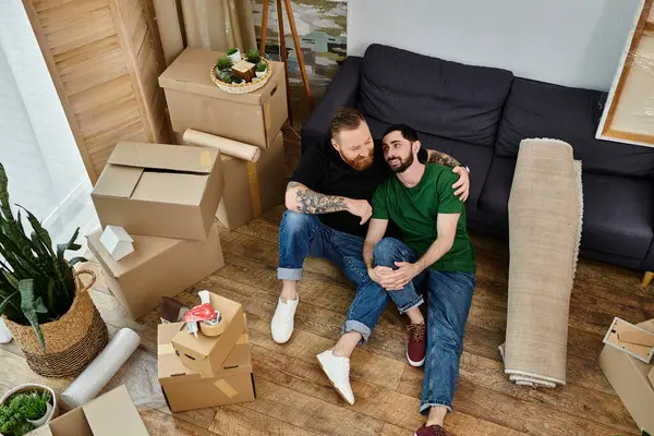 A gay couple sits atop a wooden floor, contemplating their new life together as they move into their new home. — Stock Photo