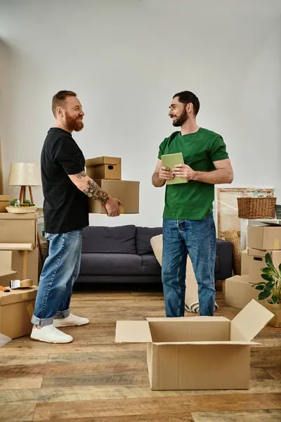 Гей-пара стоїть пліч-о-пліч у кімнаті, наповненій рухомими коробками, сигналізуючи про новий початок у своєму новому будинку. — стокове фото