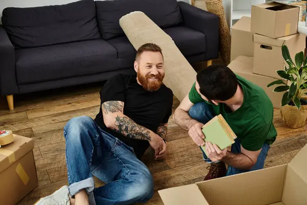 Una pareja gay se sienta en el suelo rodeada de cajas móviles, embarcándonos en un nuevo capítulo en sus vidas juntos. - foto de stock