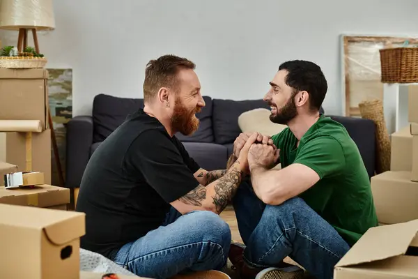 Due uomini innamorati, seduti in mezzo a scatole mobili, che si abbracciano nella loro nuova casa. — Foto stock