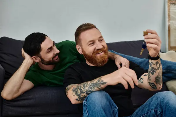 A gay couple relaxes atop a couch in their new home, taking a moment together, looking at key — Stock Photo