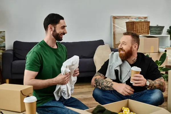 A gay couple in love takes a break, sitting atop a couch surrounded by moving boxes in their new home. — Stock Photo